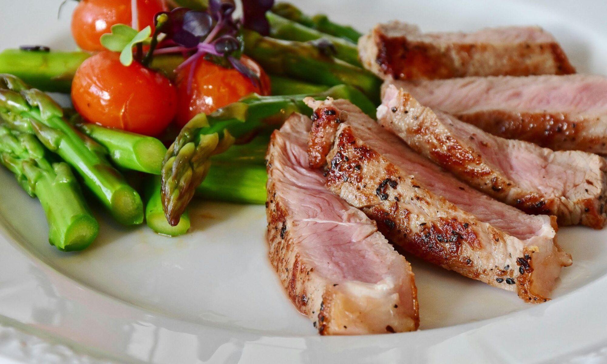 Grilled pork slices with pepper crust, served with asparagus and blistered cherry tomatoes on a white plate; greens garnish visible.